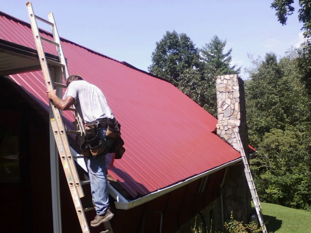 Roofer on a ladder beside a red metal roof and stone chimney — Soffit & Fascia Repair by Roofing Brothers in Evergreen